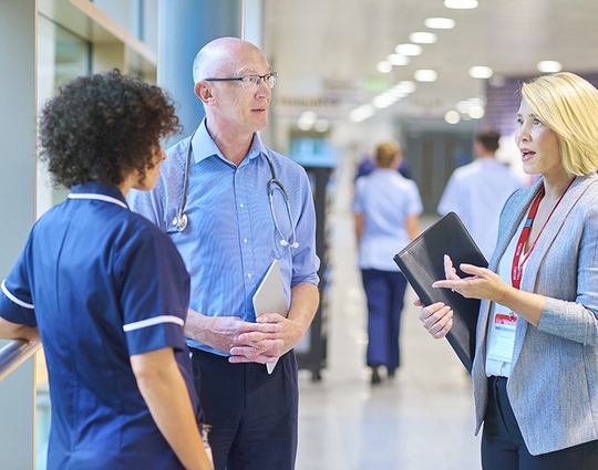 a business woman chats to a doctor and senior staff nurse in a busy hospital corridor