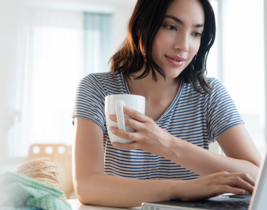A woman holding a coffee mug uses a laptop computer