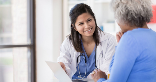 A young nurse uses laptop at a desk, while talking and smiling with a senior woman.