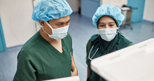Elevated view of male surgeon and female anesthetist wearing green scrubs, caps, and masks checking computer monitors in operating room.
